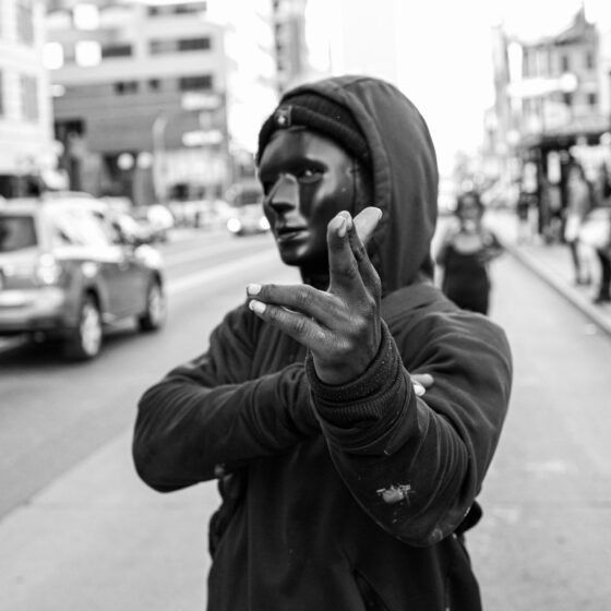 A black-and-white photo of a man with a black mask and black hood on, pointing at the camera on a city sidewalk