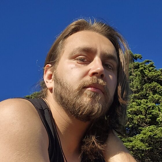 A Latino man with long, straight, blonde hair, a beard, and brown eyes. He sits outside, wearing a black tank top.