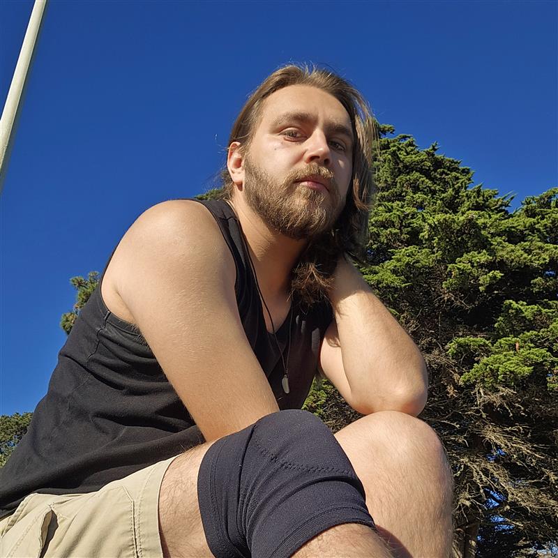 A Chicano man with medium-length hair and a beard. He is wearing a black tank top, chino shorts, and a pendant necklace.
