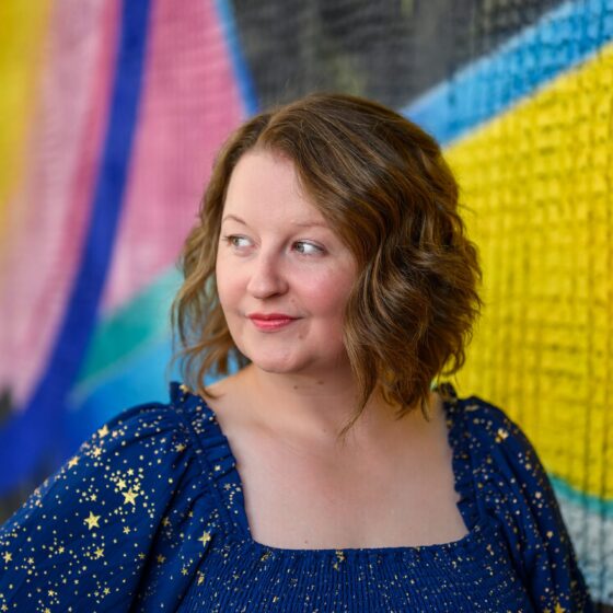 Erica Reid, a white woman with short brown hair, wears a navy blue square-necked top with puffed sleeves, a ruched, bodice, and gold stars all over. She stands in front of a multicolored mosaic and looks sideways to her right.