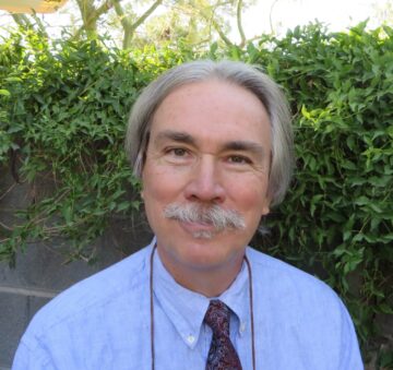 A white man with gray hair parted in the middle and reaching his ears on either side. He has a gray mustache and is wearing a light-blue shirt and red tie as he stands in front of greenery and a bit of wall.