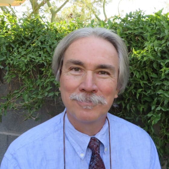 A white man with gray hair parted in the middle and reaching his ears on either side. He has a gray mustache and is wearing a light-blue shirt and red tie as he stands in front of greenery and a bit of wall.