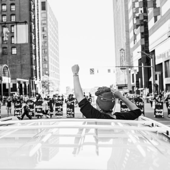 A woman holding her head and arms out of the sunroof of a car, facing a line of Black Lives Matter protesters