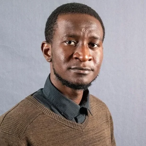 A man with buzzed black hair, a short beard, and dark-brown eyes. He stands in front of a gray background, wearing a brown sweater over a dark-gray collared shirt.