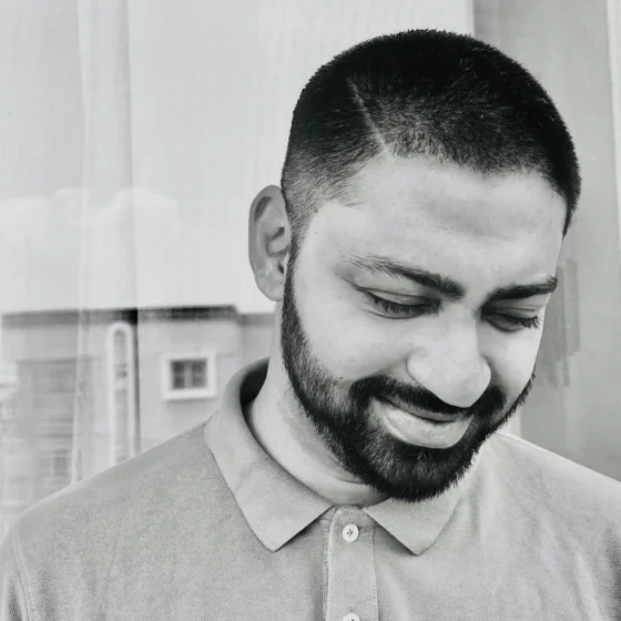 A black-and-white photo of a man with buzzed, dark hair and beard, looking down and smiling. He wears a collared, button-up shirt.