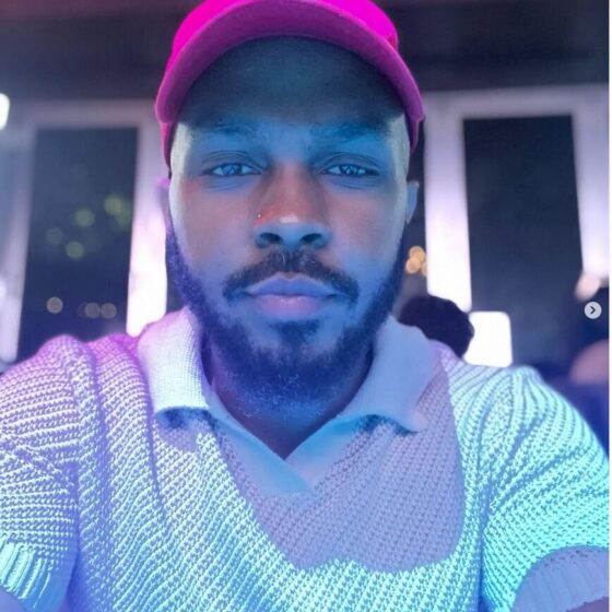 A Black man with a beard in a ballcap. He's lit by a bluish light