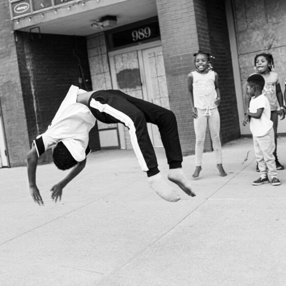 A black-and-white photo of a teenager doing a backflip on a sidewalk for a group of excited children