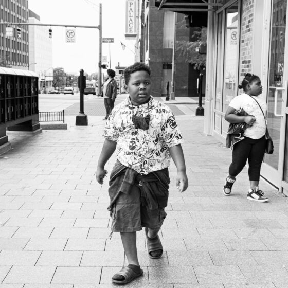 A young boy walking down a brick sidewalk in a city, wearing a printed shirt, dark cargo shorts, and slides
