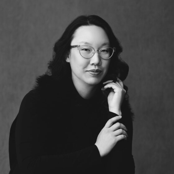 A black-and-white photo of a woman with black, wavy hair. She wears a black, long-sleeved shirt and cat-eye glasses