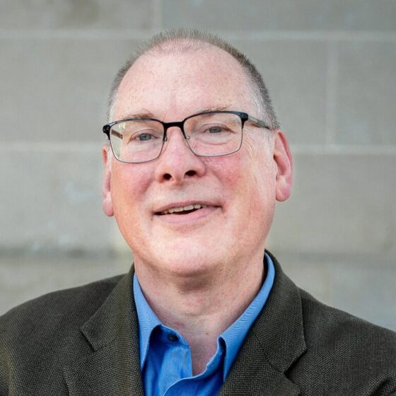 A man with buzzed, gray hair and blue eyes, smiling. He wears a blue, collard shirt underneath a dark-gray blazer