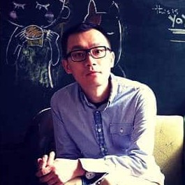 A man with short, black hair and brown eyes. He sits in front of a chalkboard, wearing a light-blue, button-down shirt.