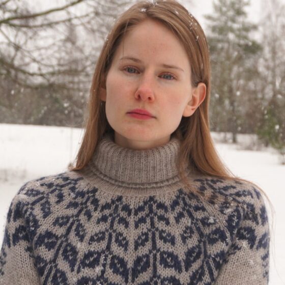 A woman with long, straight, strawberry-blonde hair and blue eyes. She stands outside in the snow, wearing a gray-and-blue, wool sweater.