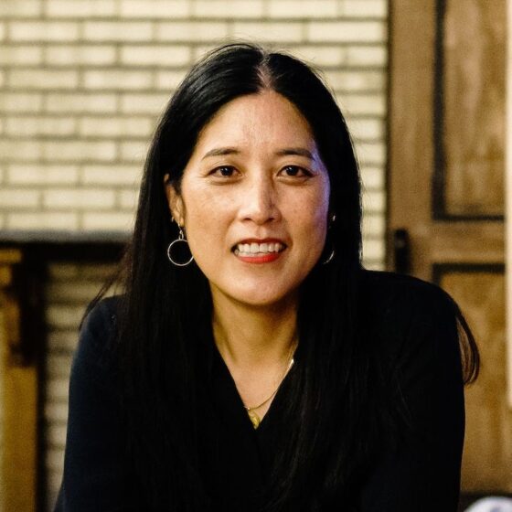 A woman with long, straight, black hair and brown eyes, smiling. She wears a black, long-sleeved shirt