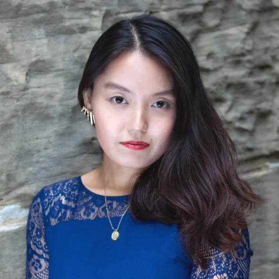A woman with long, straight, black hair and dark-brown eyes. She stands in front of a stone wall, wearing a royal-blue dress and a gold necklace