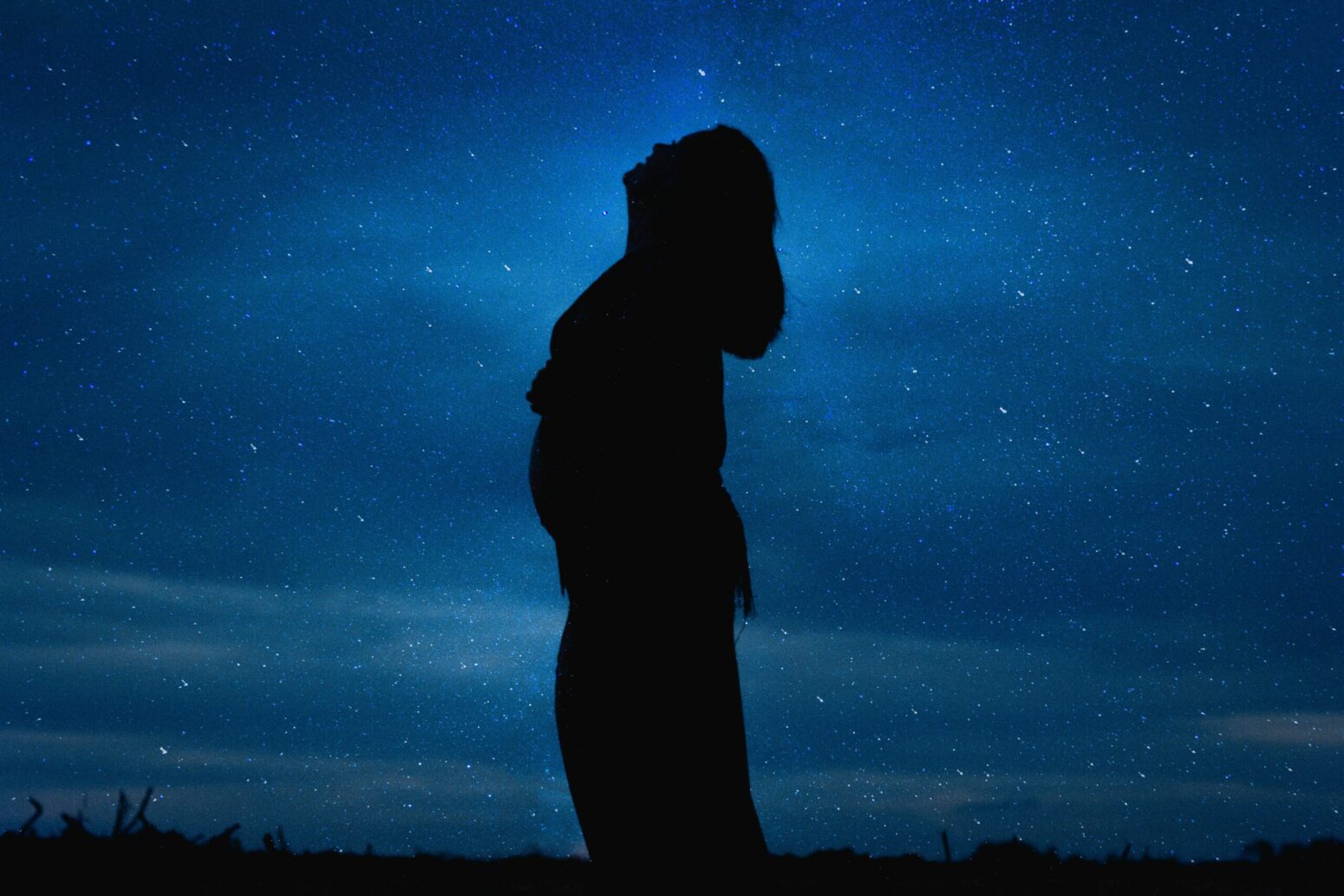 The silhouette of a pregnant woman looking up against a blue, starry sky.