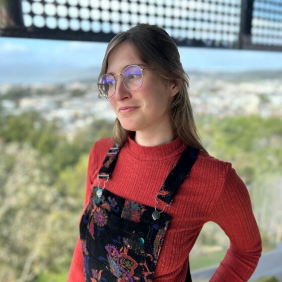 A White woman with long, straight, brown hair and green eyes. She stands near a beach, wearing a long-sleeved, red shirt, black overalls with flowers printed on them, and round glasses.