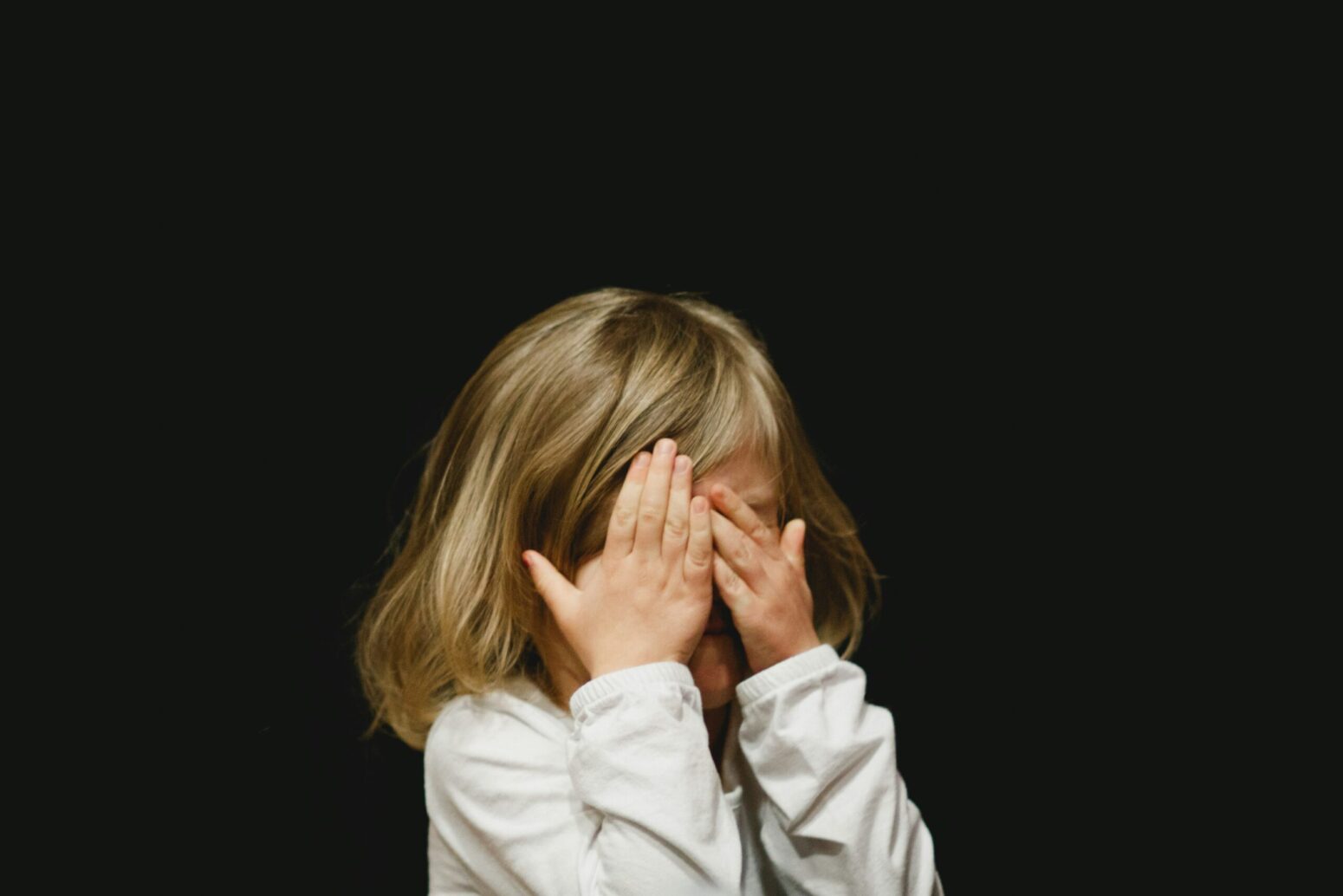A little girl with straight, blonde hair covering her face with her hands. She stands in front of a black background and wears a white shirt.