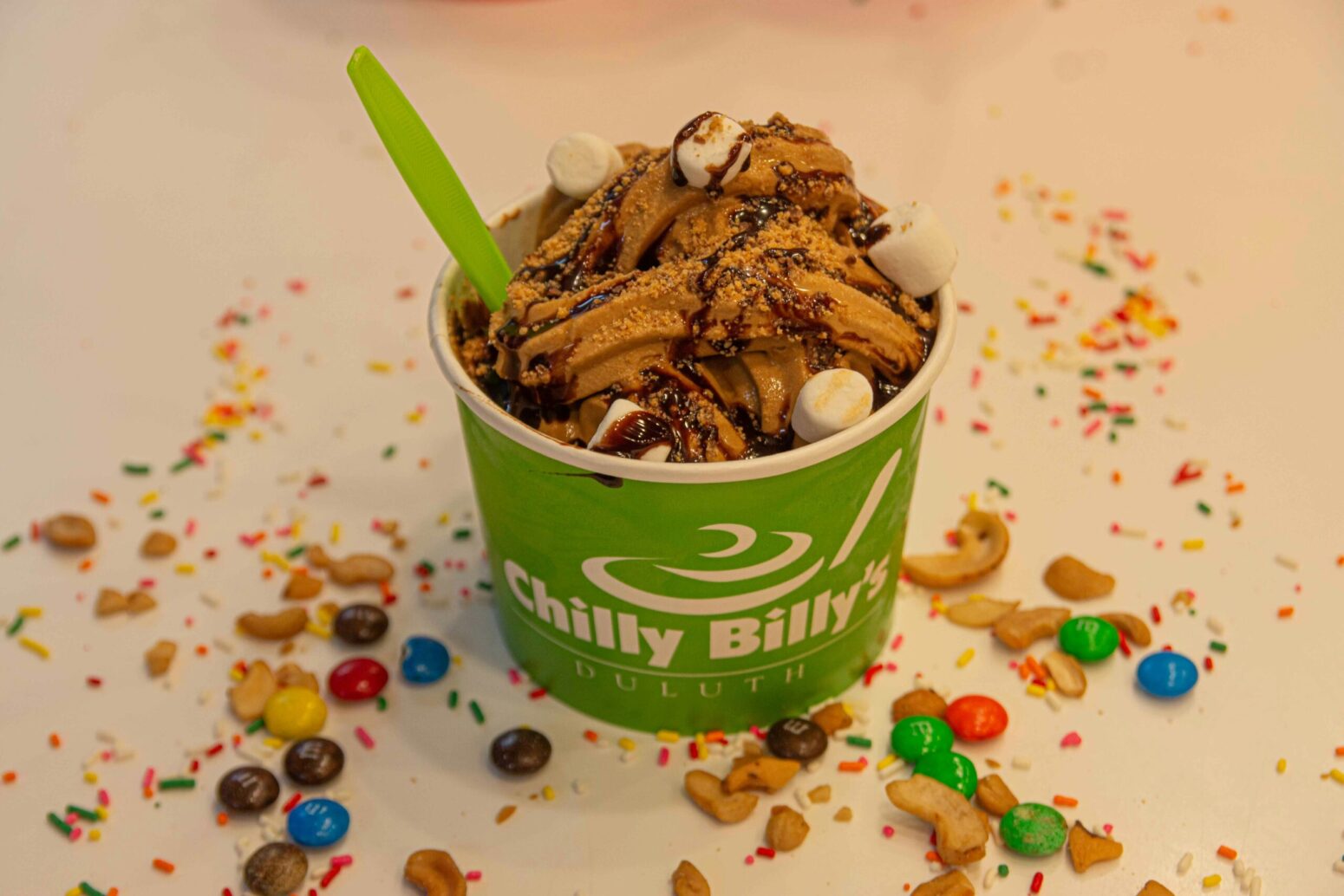 A green cup of chocolate, frozen yogurt with chocolate syrup and marshmallows on top and M&Ms surrounding it