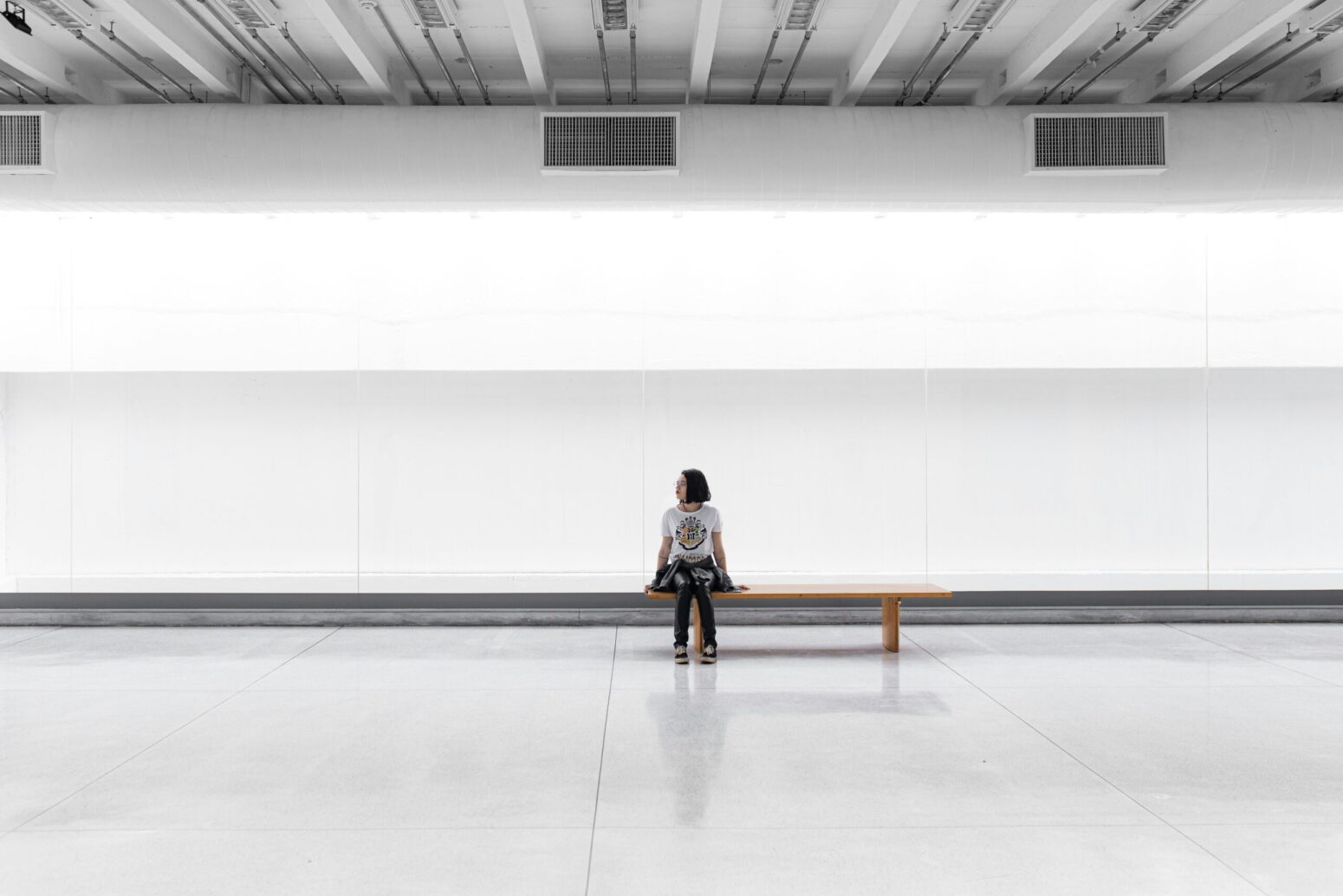 A woman sitting on a lone bench in a large, white room, looking around expectantly
