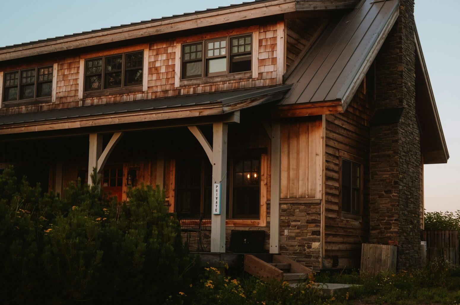 A wooden house at dusk, with a covered porch, a stone chimney, and flower bushes sitting outside