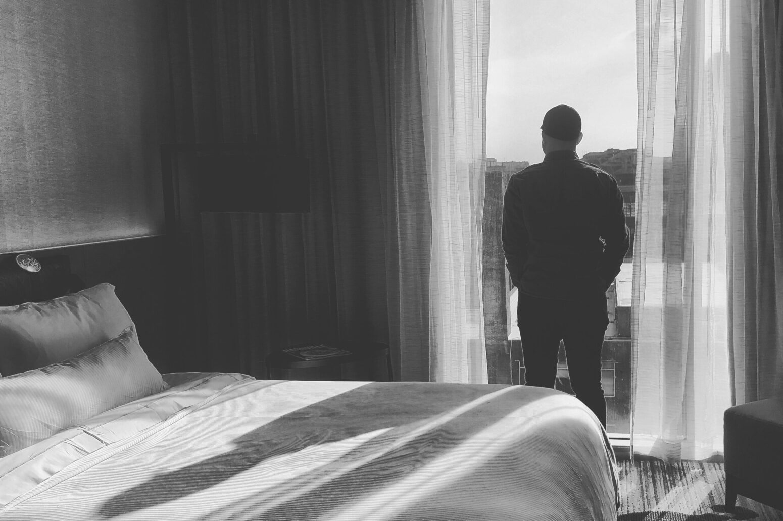 A black-and-white photo of a man looking out a tall window next to a bed