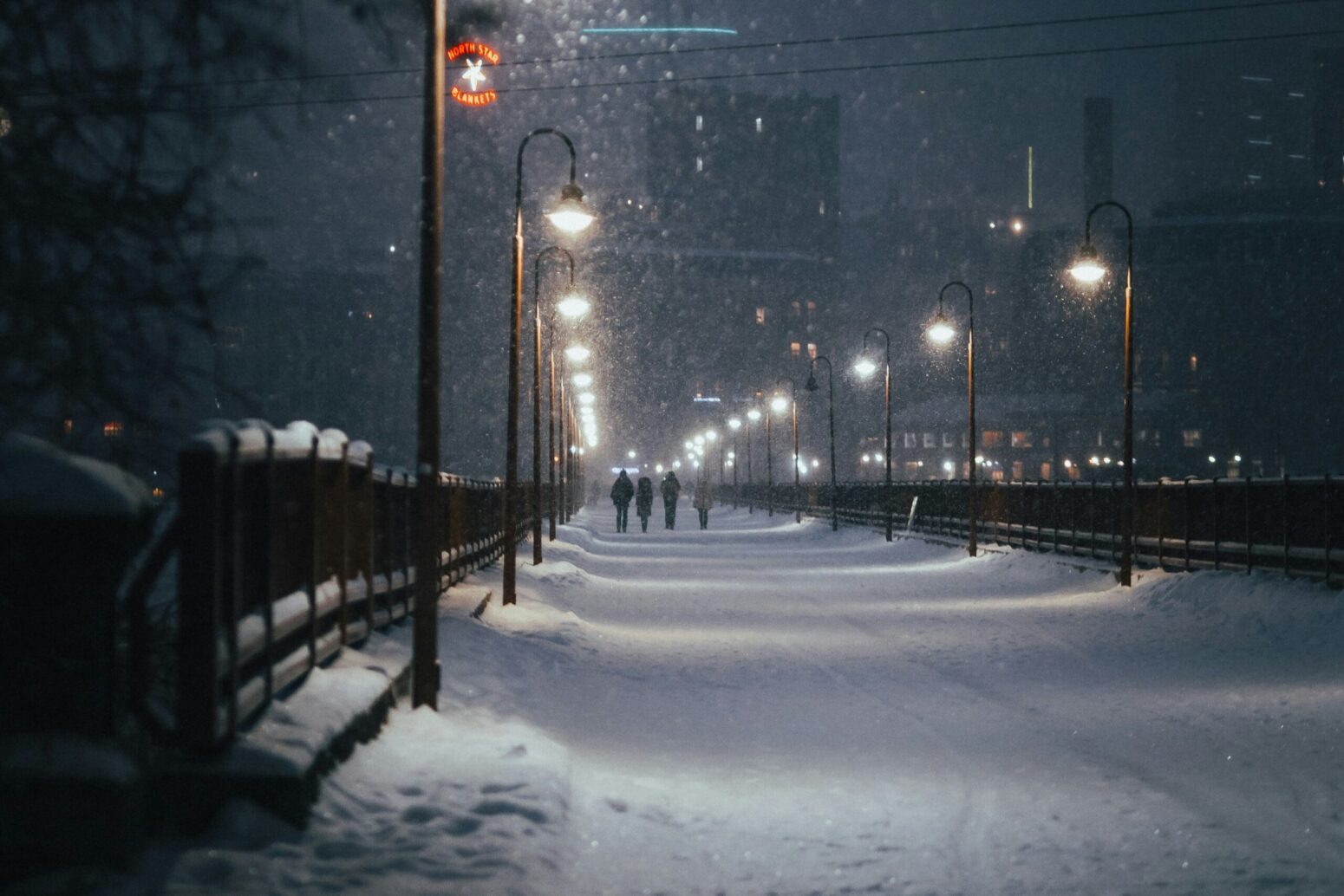 A snowy walkway at night, lit by street lamps. Four people walk side by side, a little ways down, toward a city