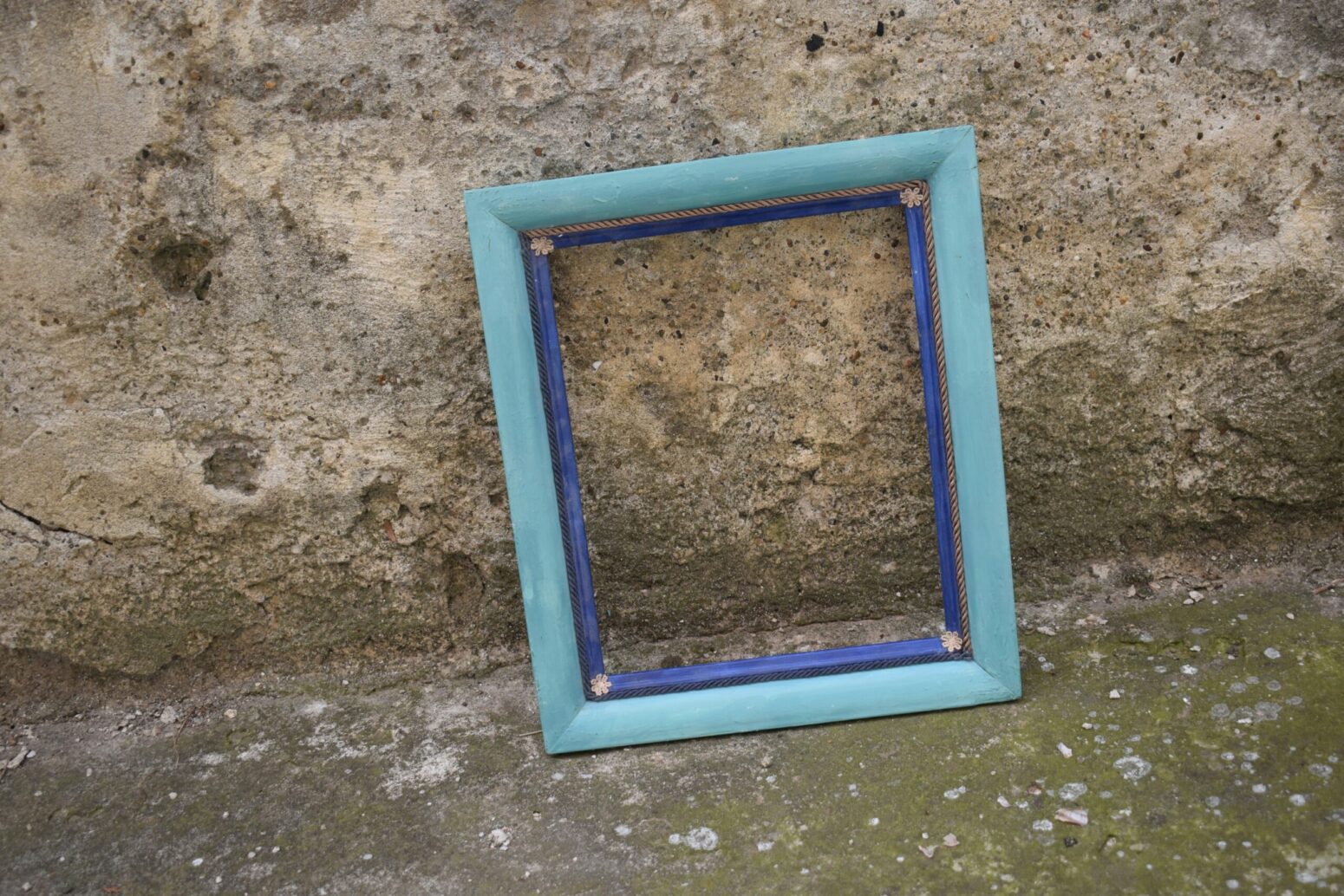 A light-blue wooden picture frame that's empty and sitting on the ground near a stone wall