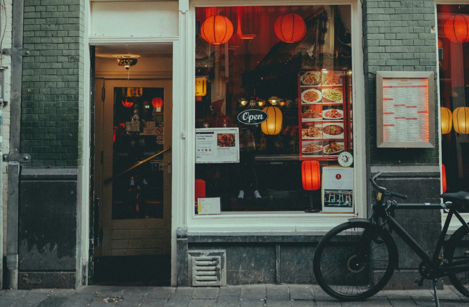 The storefront of a Chinese restaurant in an urban area. The window displays a menu, and the inside is lit with red lanterns