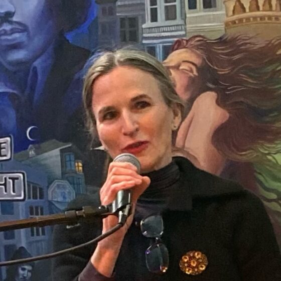 A White woman with straight, gray-blonde hair in a ponytail and brown eyes. She stands at a podium, speaking into a microphone, wearing a black, collared shirt.