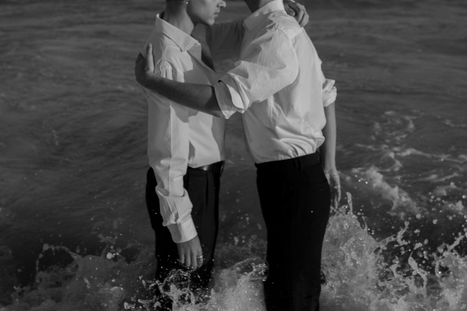 A black-and-white photo of two men standing in the ocean, embracing, and wearing white dress shirts and black pants.