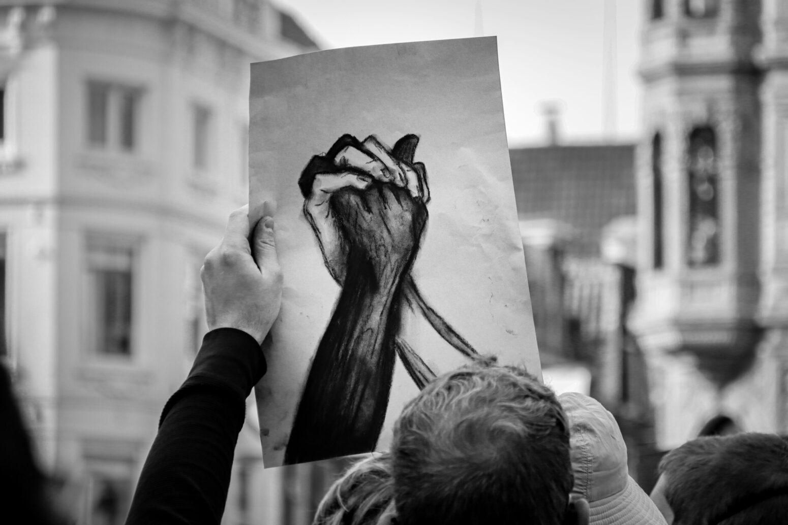 A black-and-white photo of a person holding up a charcoal drawing of two hands clasped together--one black, one white--at a protest