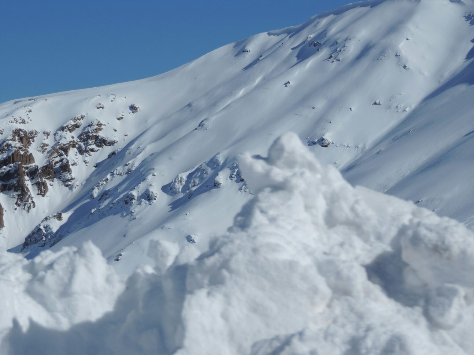 The side of a mountain, covered in snow, against a blue sky.