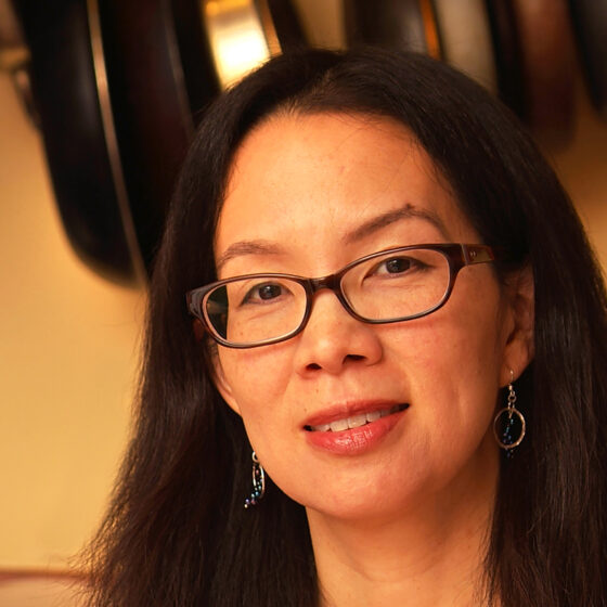 Headshot of an Asian American woman with glasses and dangling earrings. She's in front of a warm-yellow background