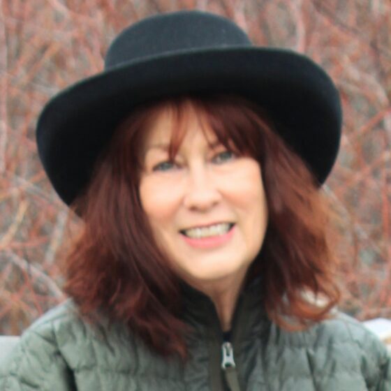 A white woman with red hair, wearing a broad-rimmed black hat and a green jacket. She's sitting outside with bare-branched trees behind her