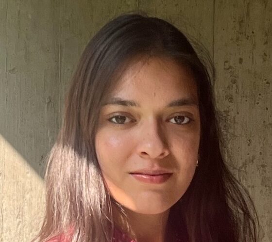 A Southeast Asian woman with long dark hair slightly past her shoulders. She's wearing a deep pink shirt and standing partly in sun, partly in shadow, with a rock or concrete wall behind her