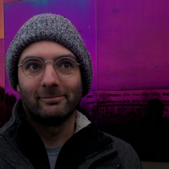 A man with Pakistani and white ancestry looks up and to the right. He's wearing glasses and a gray knit cap and wearing a dark coat, in front of a purplish screen