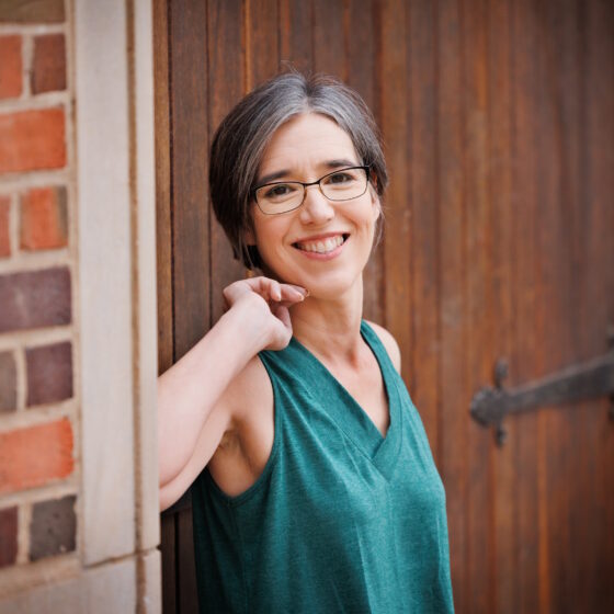 A white woman with black and gray hair in a short pixie cut. She's wearing glasses and a teal sleeveless shirt and leaning on a wide wooden door next to a brick wall.