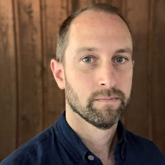 Fritz Horstman, a middle-aged white man with a short beard and mustache, stands in front of a wooden wall. He wears a blue button-down and doesn't smile.