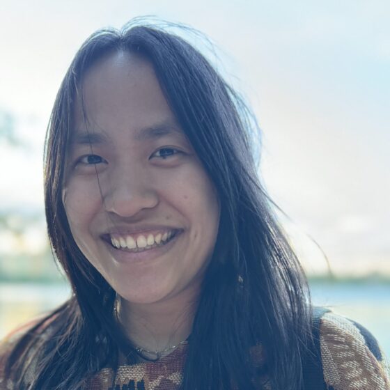 An Asian American woman with long black hair, smiling widely. She's wearing a multicolored shirt and standing outside near water, but her face is in shadow.