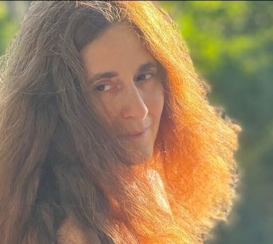 A white woman with brown wavy hair blowing in the wind. She is looking off to the left, and her left side has sunlight on it.
