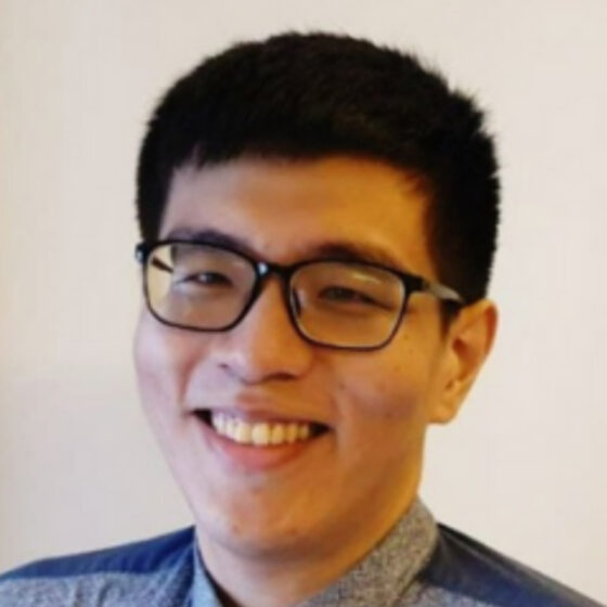 Head shot of an Asian man with glasses and short dark hair. He's smiling, wearing a gray-and-blue shirt, and standing in front of a neutrally colored wall