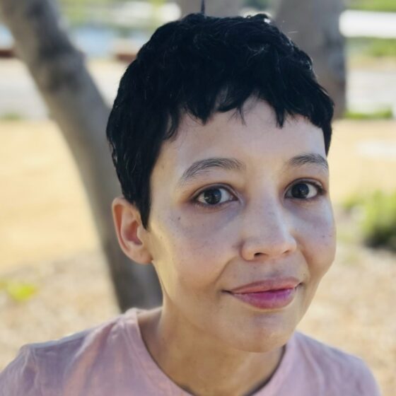 A woman with dark shortly cropped hair. She's wearing a light-pink T-shirt and leaning toward the camera as she stands outside near a tree