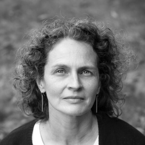 A black-and-white photo of a white woman in a white-appearing blouse and dark sweater. She has curly hair to her shoulders and dangling earrings.