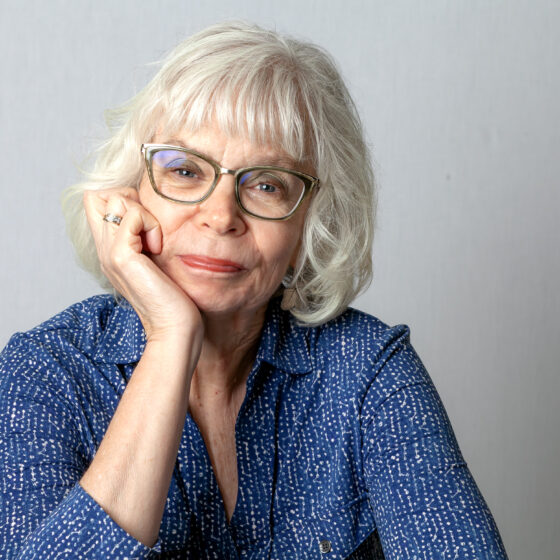 An older white woman with a blue textured top on. She has white hair and glasses and is smiling and resting her face on her right hand.