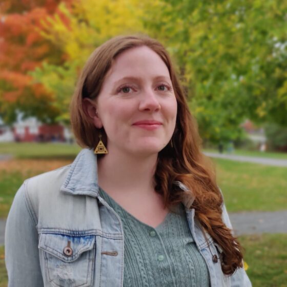 A white woman with red hair. She's standing outside with fall-changing trees behind her, and she's wearing a jean jacket and neutral-colored shirt