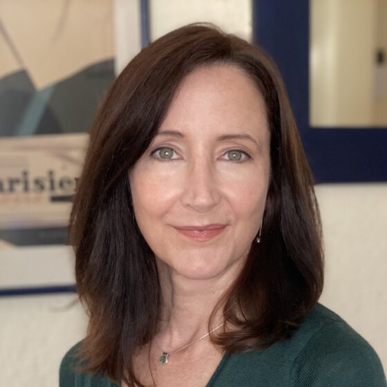 A white woman with dark hair to her shoulders and blue eyes. She's wearing a necklace and a dark gray shirt and standing in front of framed art