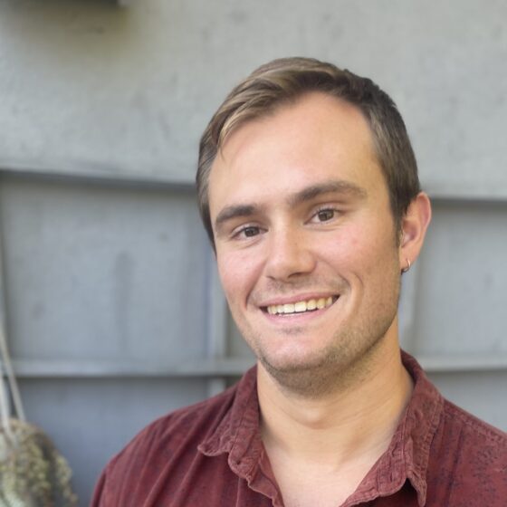 A man with light brown hair and a maroon collared shirt stands in front of gray siding and a hanging plant