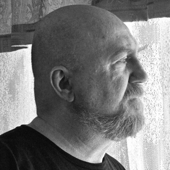 Black-and-white photo of a white man in profile, looking toward the right of the image. He is bald and has a beard and is standing in front of a lacy curtain