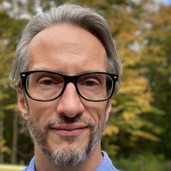 A white man with gray hair and beard. He's wearing black glasses and a blue collared shirt and standing outside with trees behind him