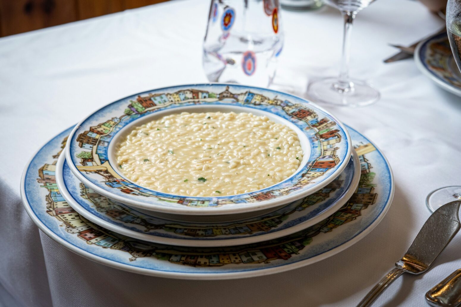 A bowl of watery risotto with some herbs in a pattern of china, which the same as the two plates below it. There's a white tablecloth on the table, silverware to the right, and a water and wine glass in the background